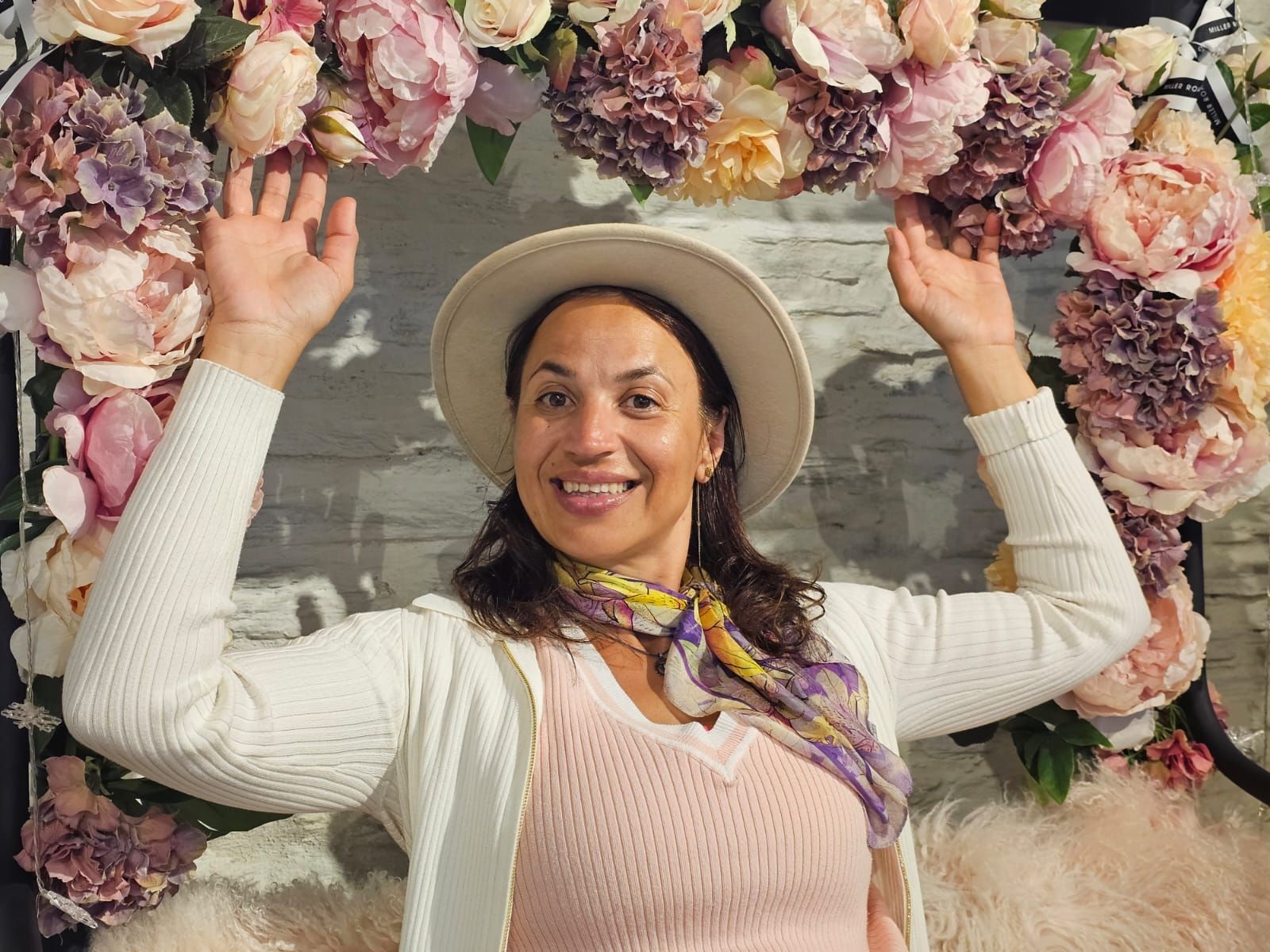 Woman in a hat smiling, standing under an arch adorned with pink and purple flowers.